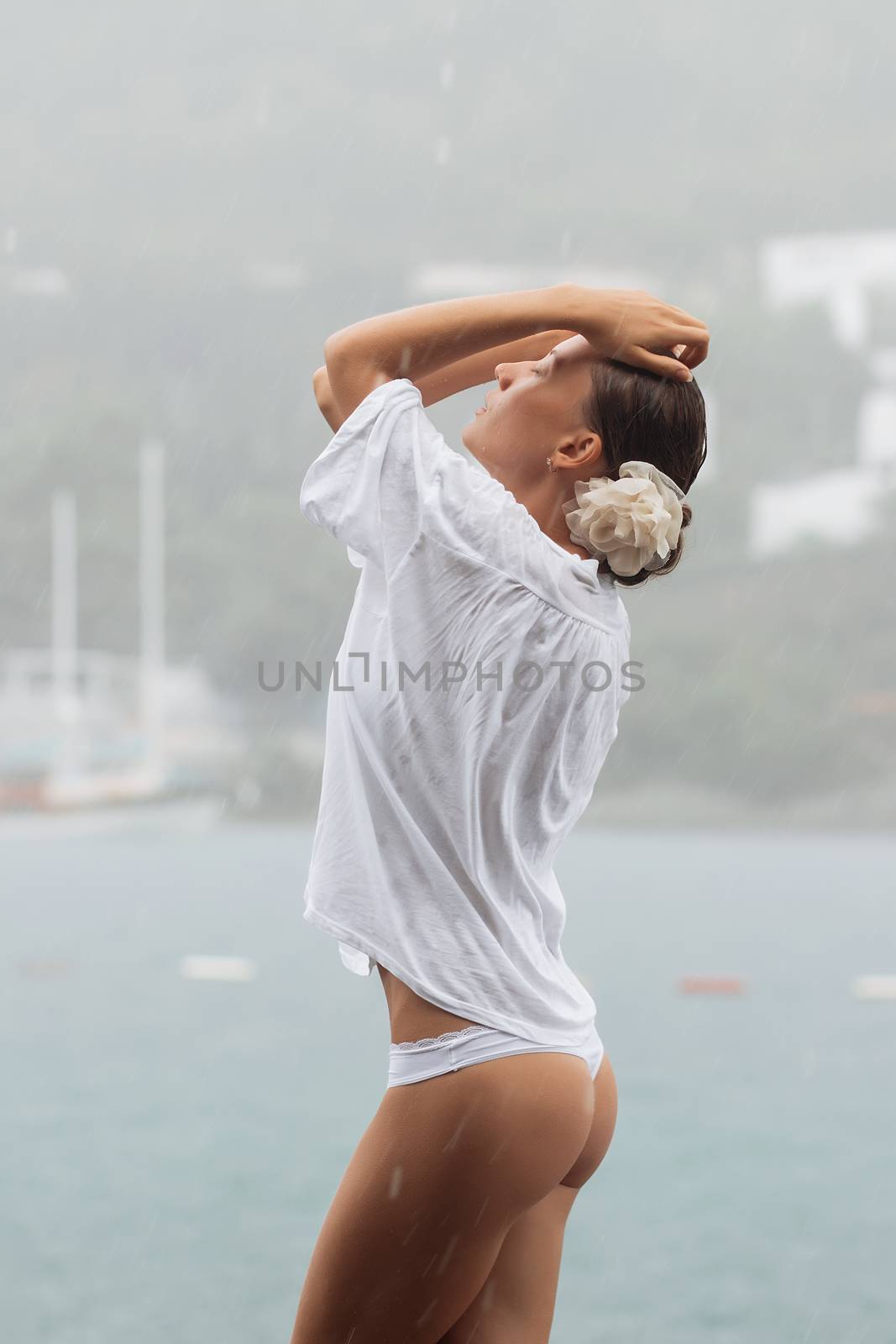 Attractive young female in while blouse and panties embracing herself and keeping eyes closed while standing near water on windy day