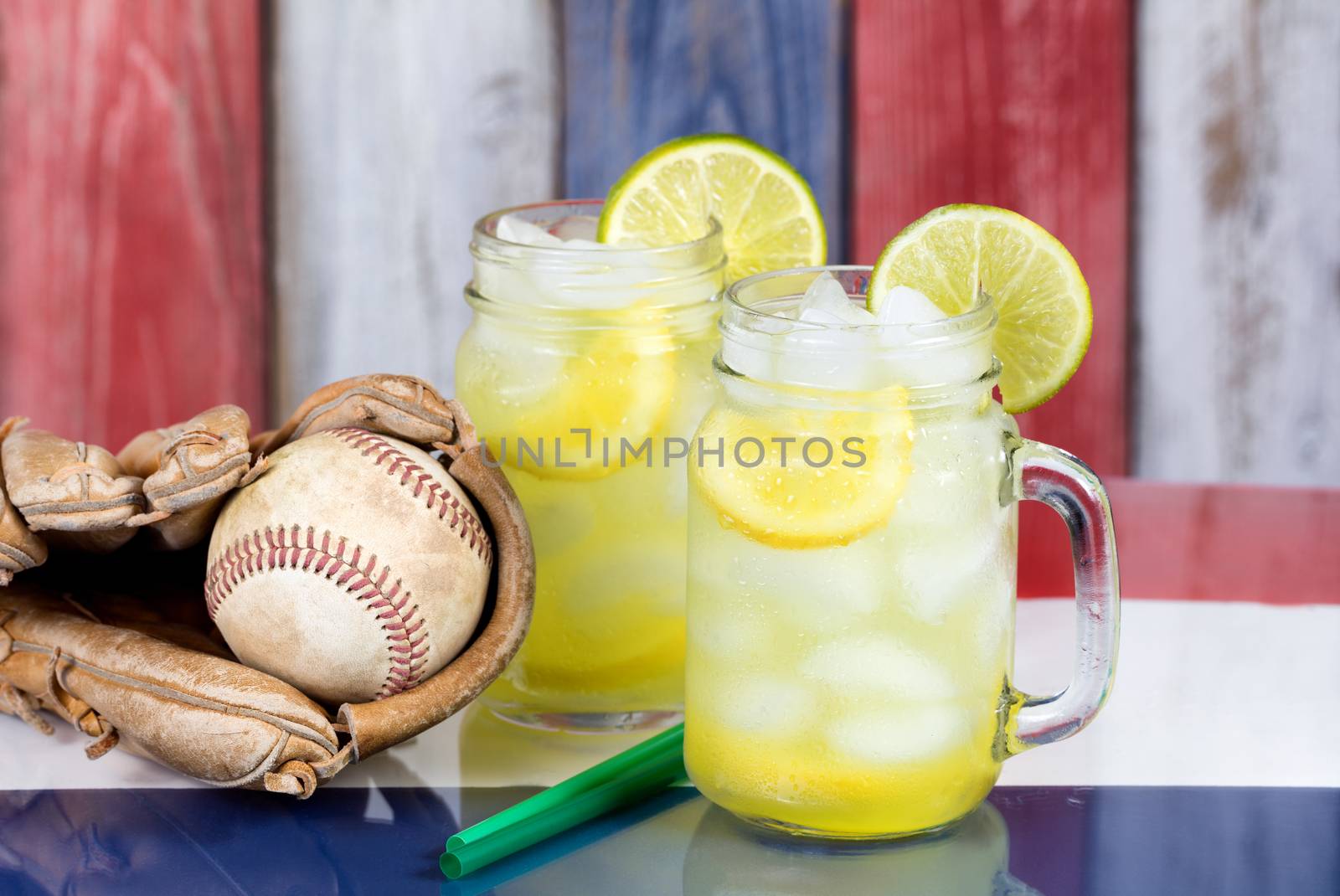 Close up of a glasses filled with cold lemonade with baseball glove and ball.  Faded wooden boards painted red, white and blue in background. Selective focus on upper front jar glass with lime slice. 