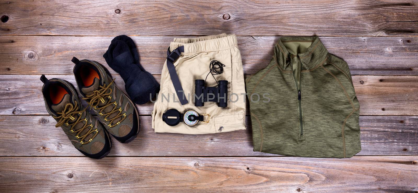 Top view of hiking gear to include hiking shoes, shorts, shirt, compass, socks and binoculars on rustic wooden boards