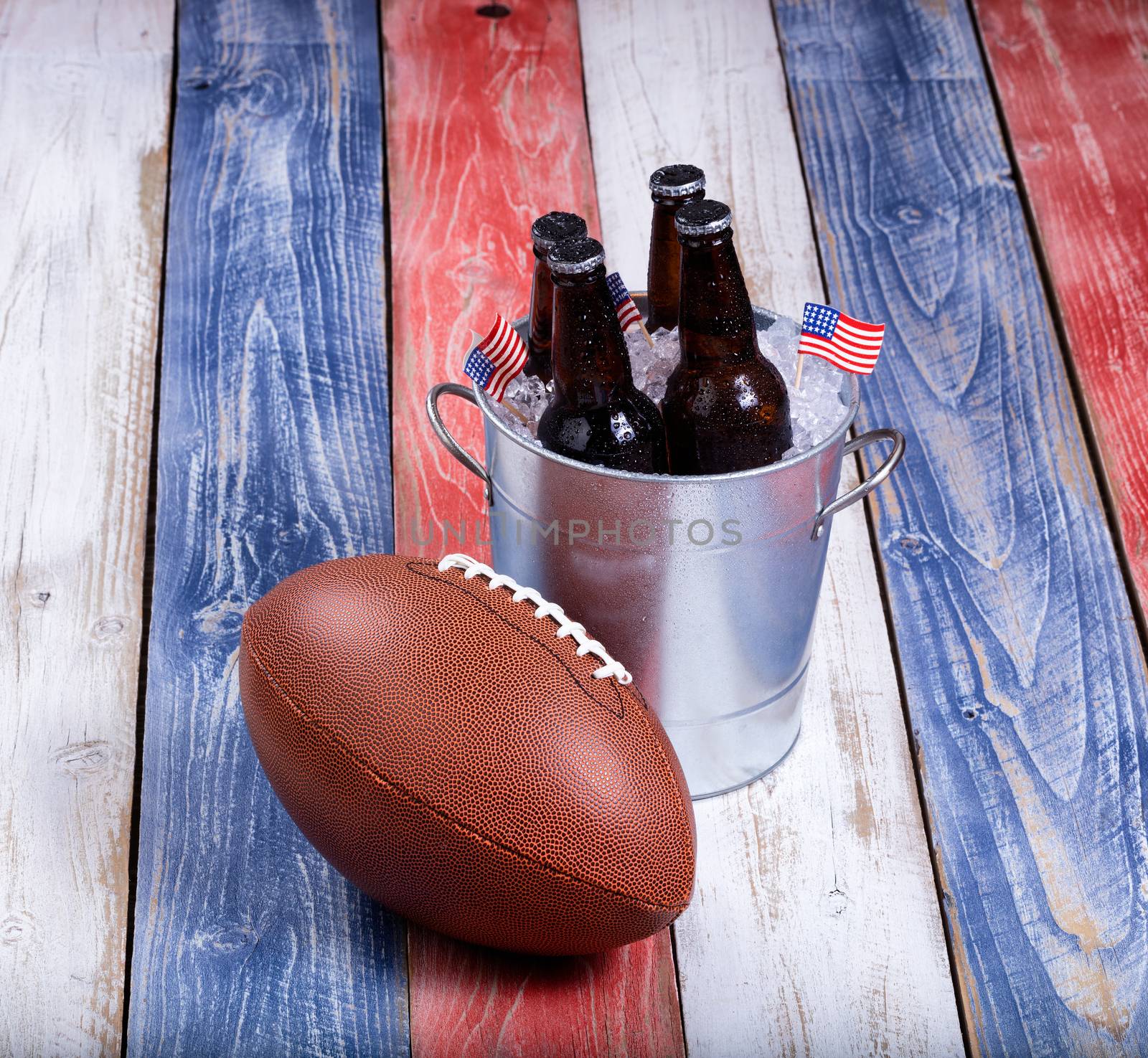 Top view of American football and bucket of ice cold beer on rustic wooden boards painted in USA national colors.