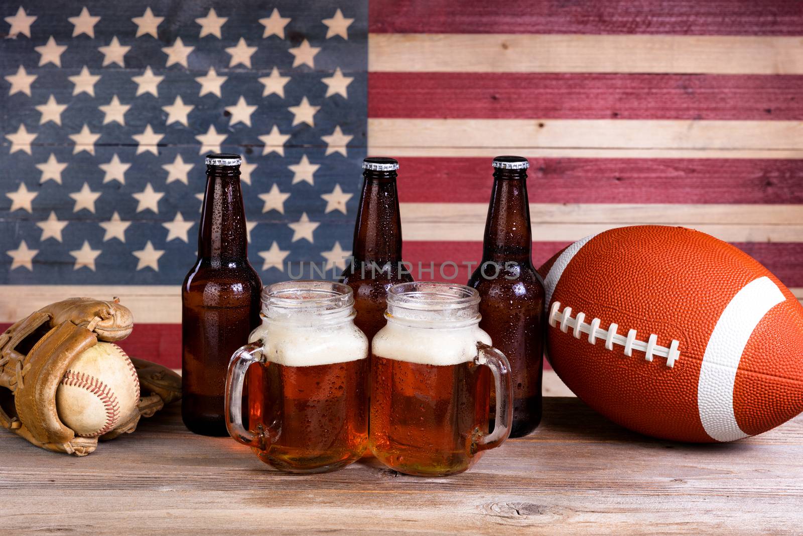 Two pint jars filled with beer, full bottles, football and baseball mitt with vintage wooden USA flag in background.