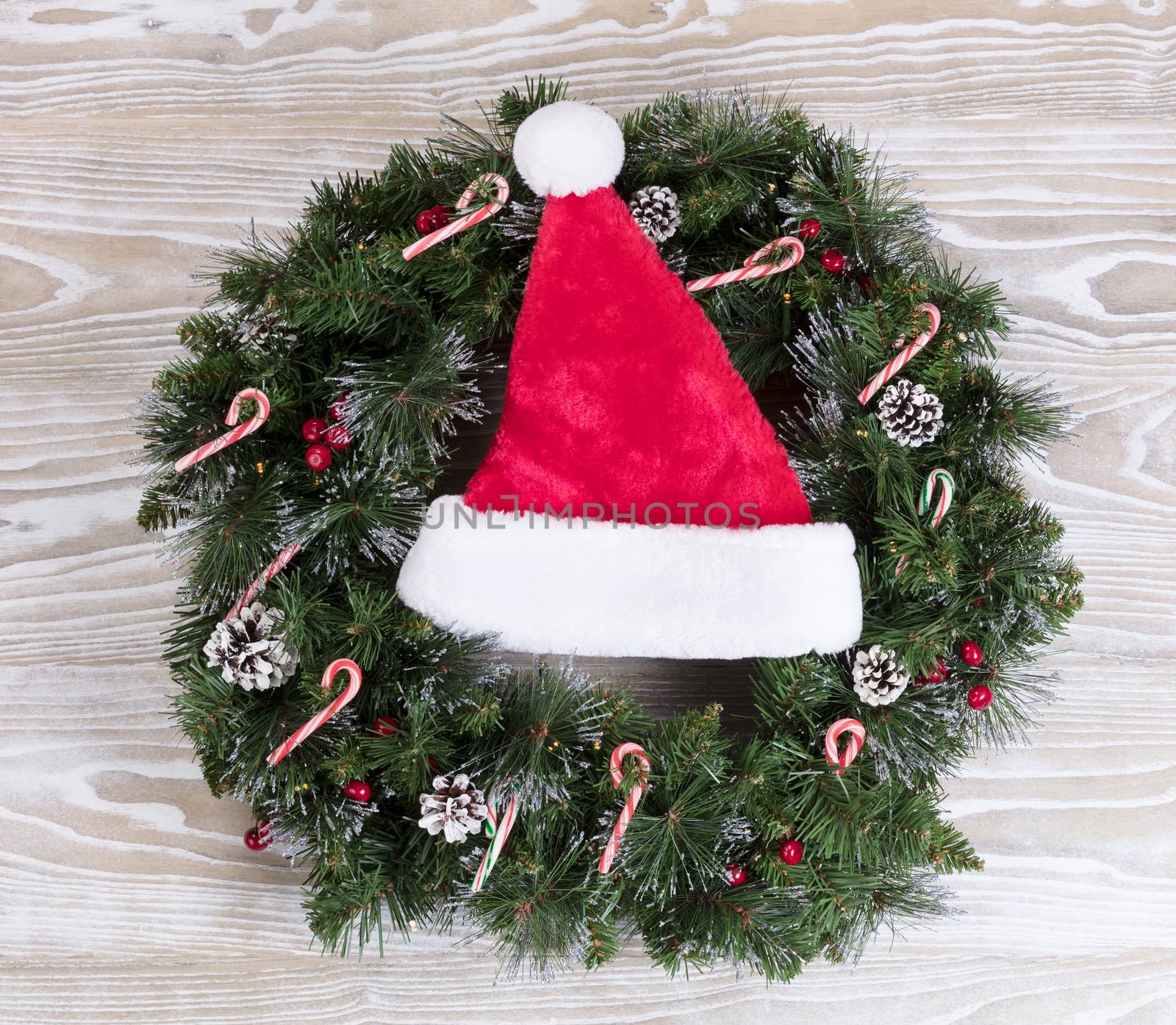 Evergreen Christmas wreath with Santa cap, lights, pine cones and candy canes on white wood. 