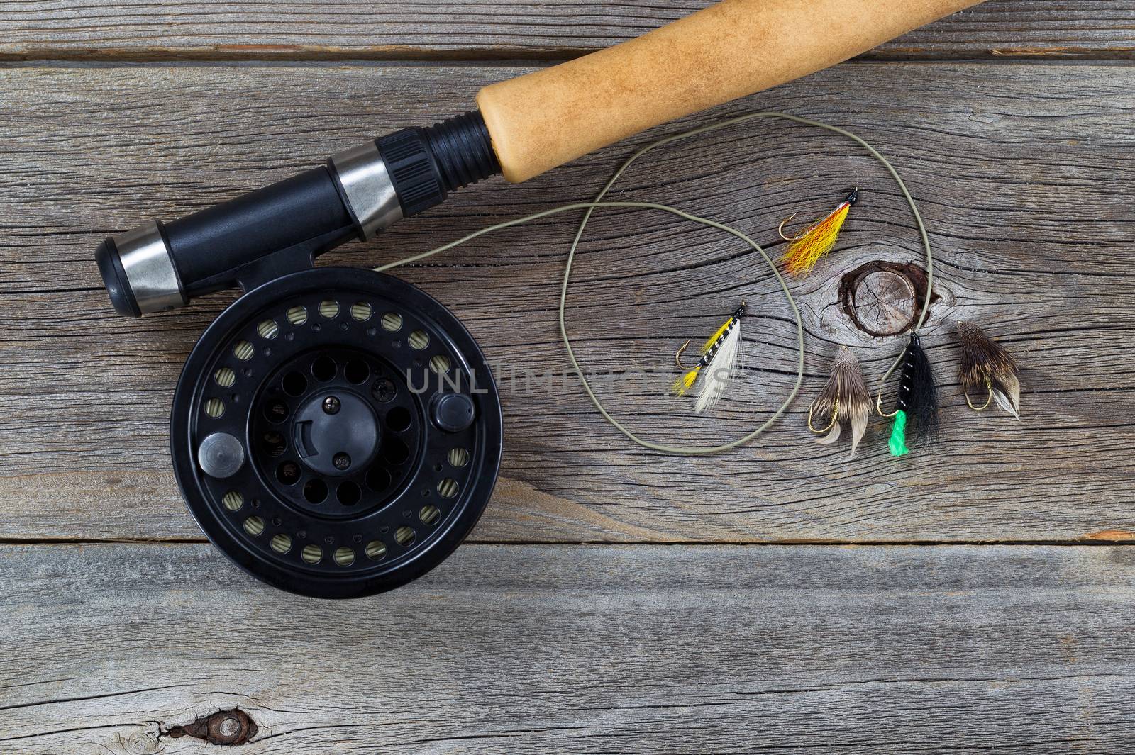 Close up of a fly reel, with line and assorted flies, and partial cork handled pole on rustic wooden boards 