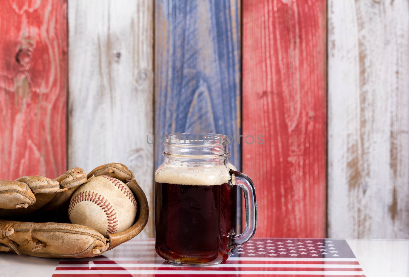Front view of a glass of cold dark beer with a worn baseball mitt and ball.  Faded wooden boards painted red, white and blue in background with USA flag underneath.  