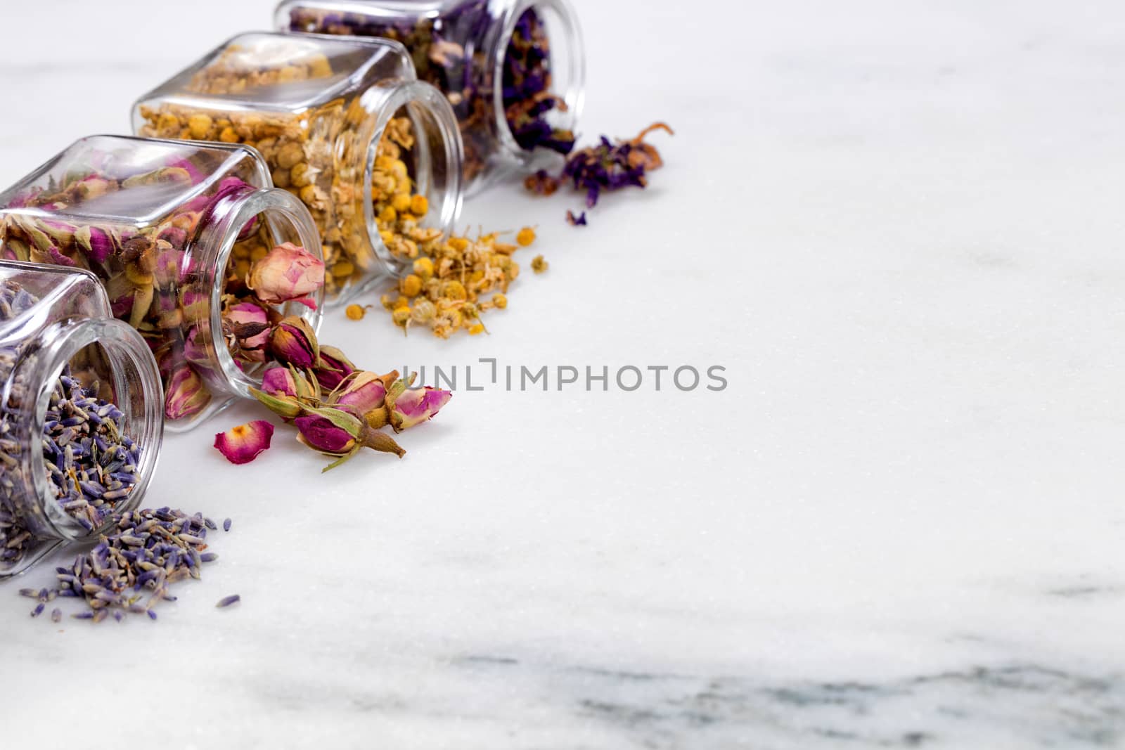 Front view of herbs and spices in glass jars spilling onto white marble stone. Selective focus on second jar with rose buds. 
