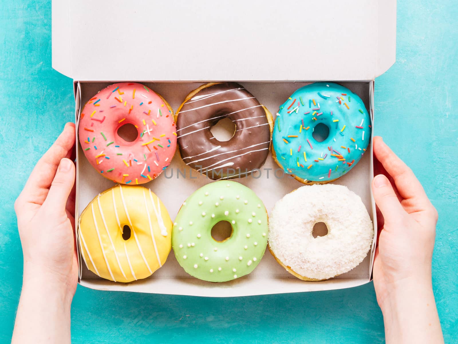 Top view of female hands holding box with colorful donuts on blue concrete background