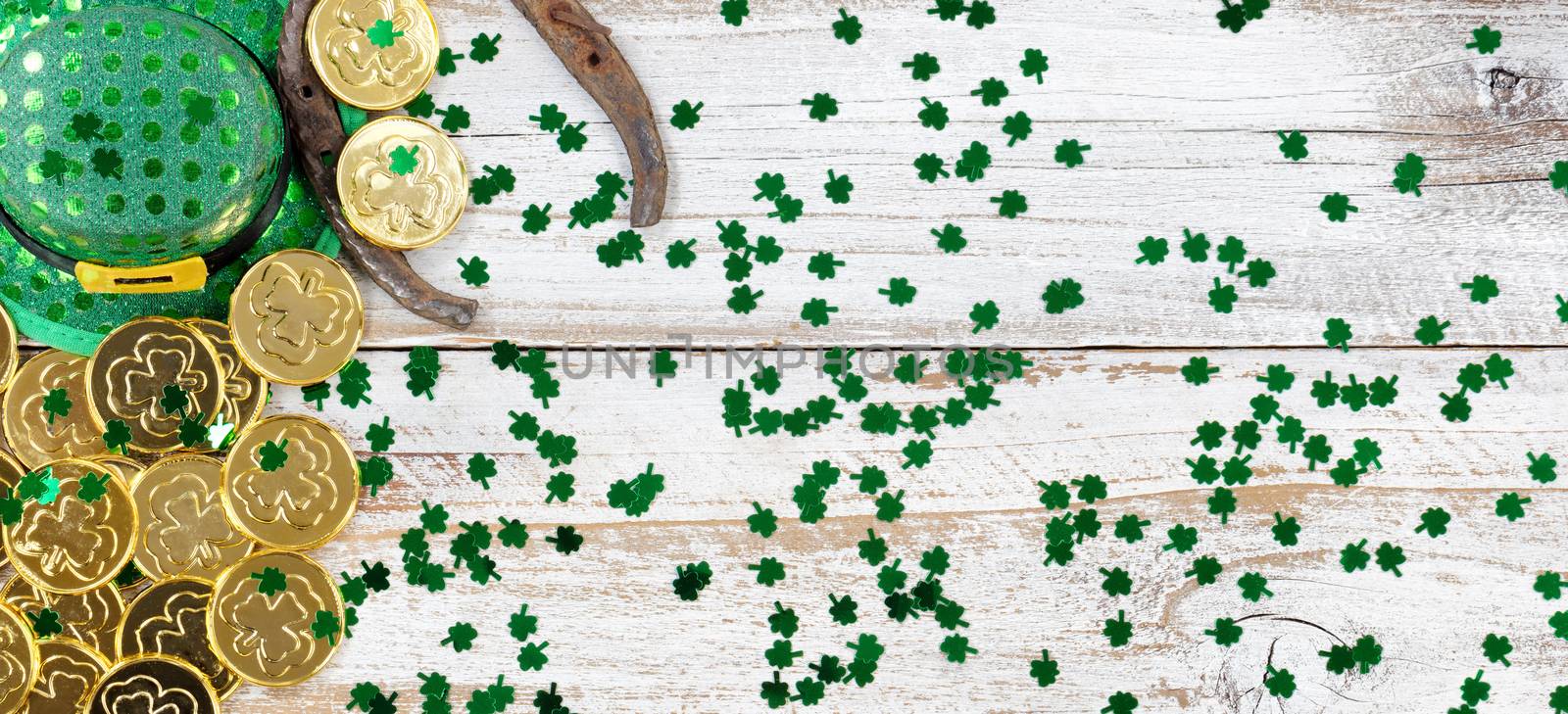 St Patrick day good luck hat, clovers and horseshoe with shiny gold coins forming left border on rustic white wooden boards 