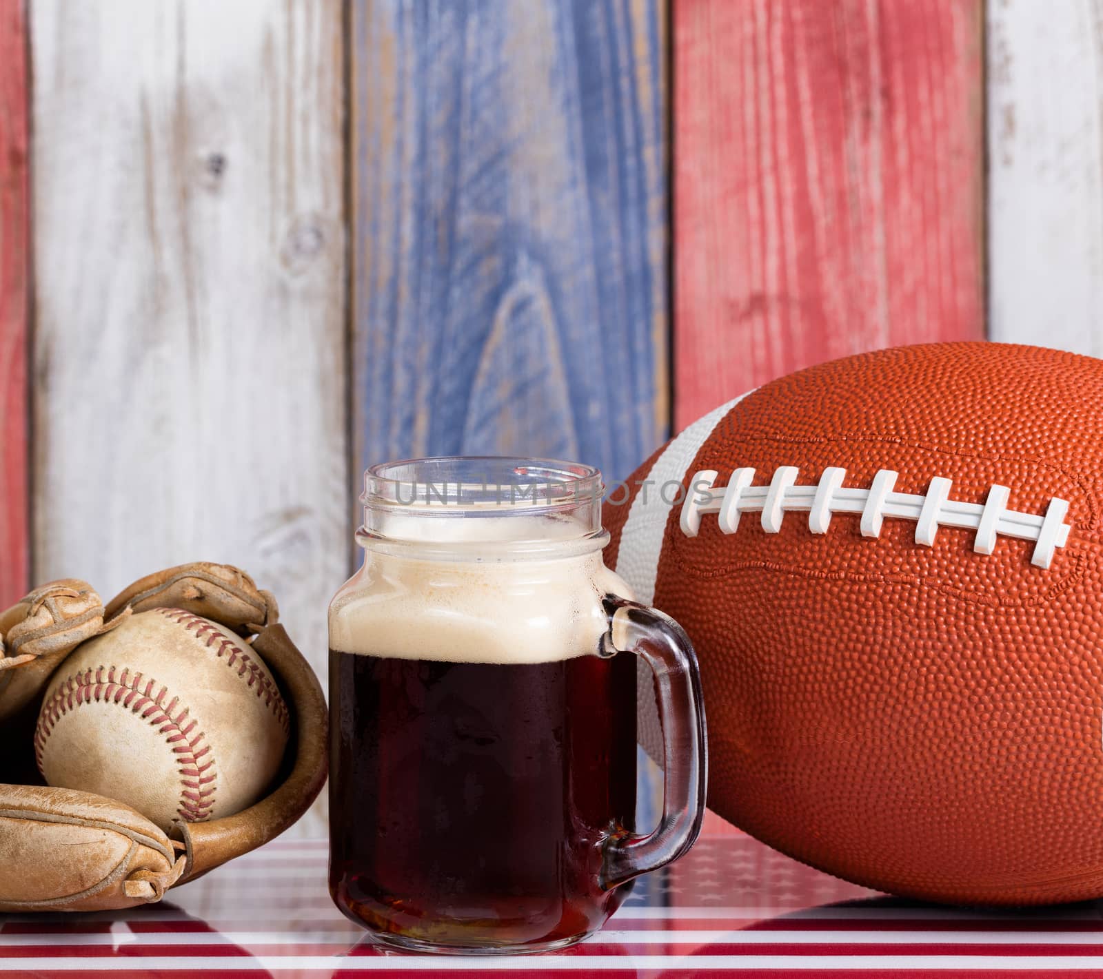 Close up front view of a glass of cold dark beer with a worn baseball mitt, ball and football.  Faded wooden boards painted red, white and blue in background with USA flag underneath.  