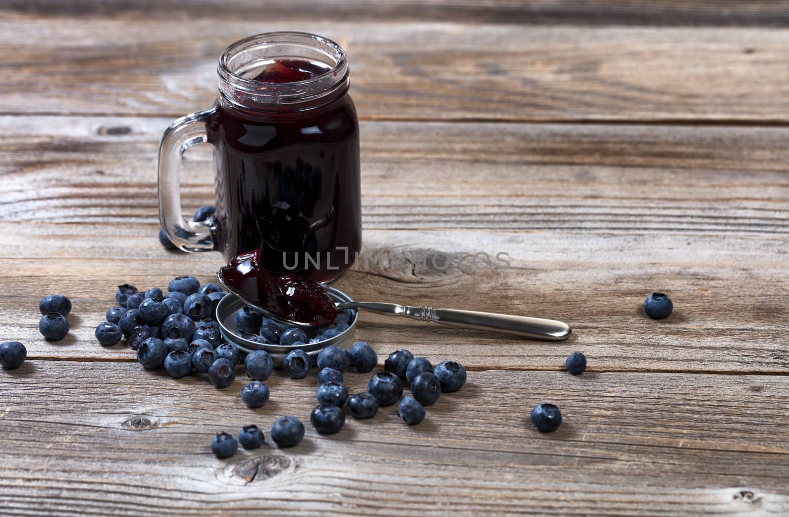 Blueberry jam in spoon with fresh ripe blueberries and jar in background on rustic wood