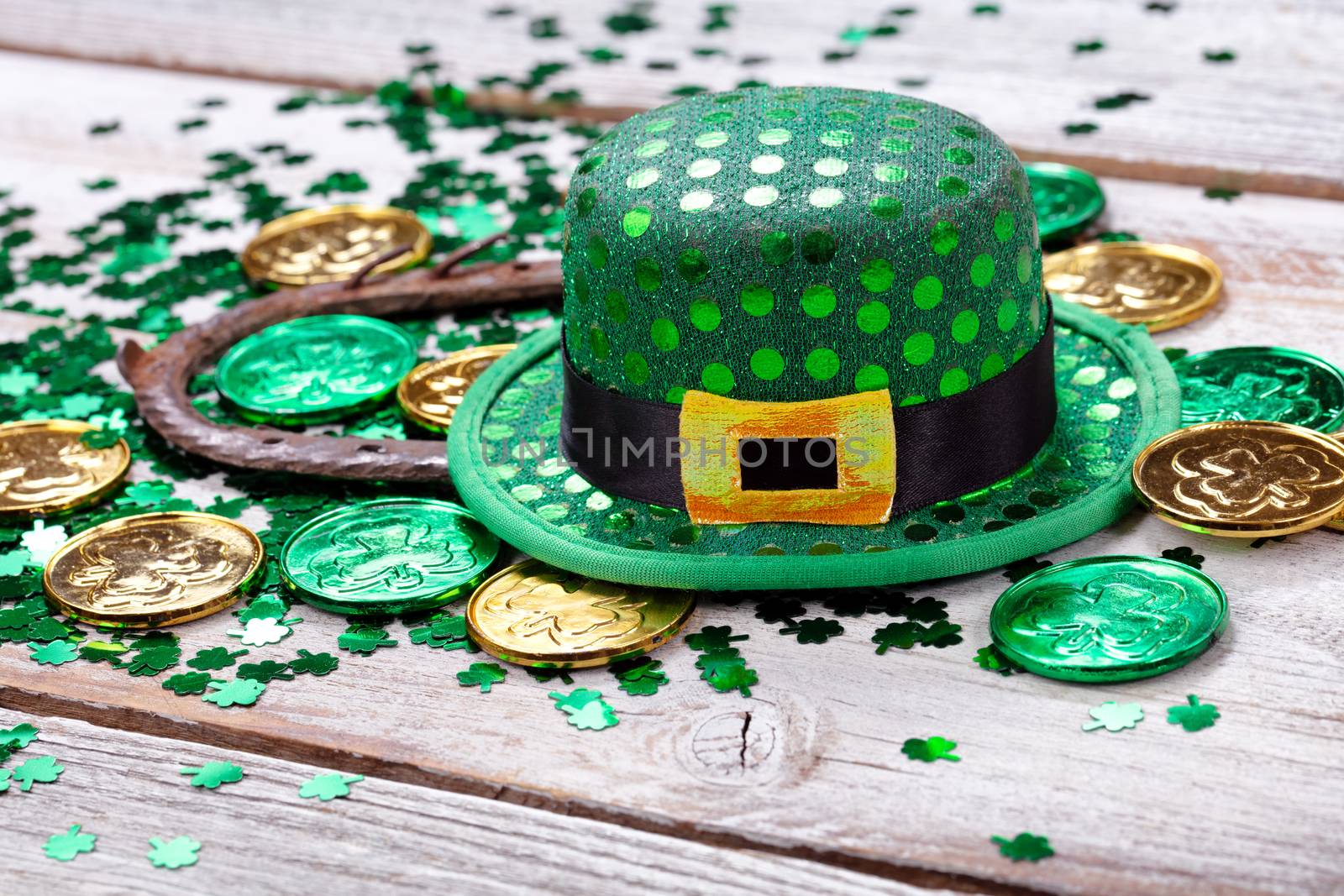 Bright green hat with gold coins, shamrock and rusty horseshoe for Saint Patrick Day on white rustic wooden background in close up format  