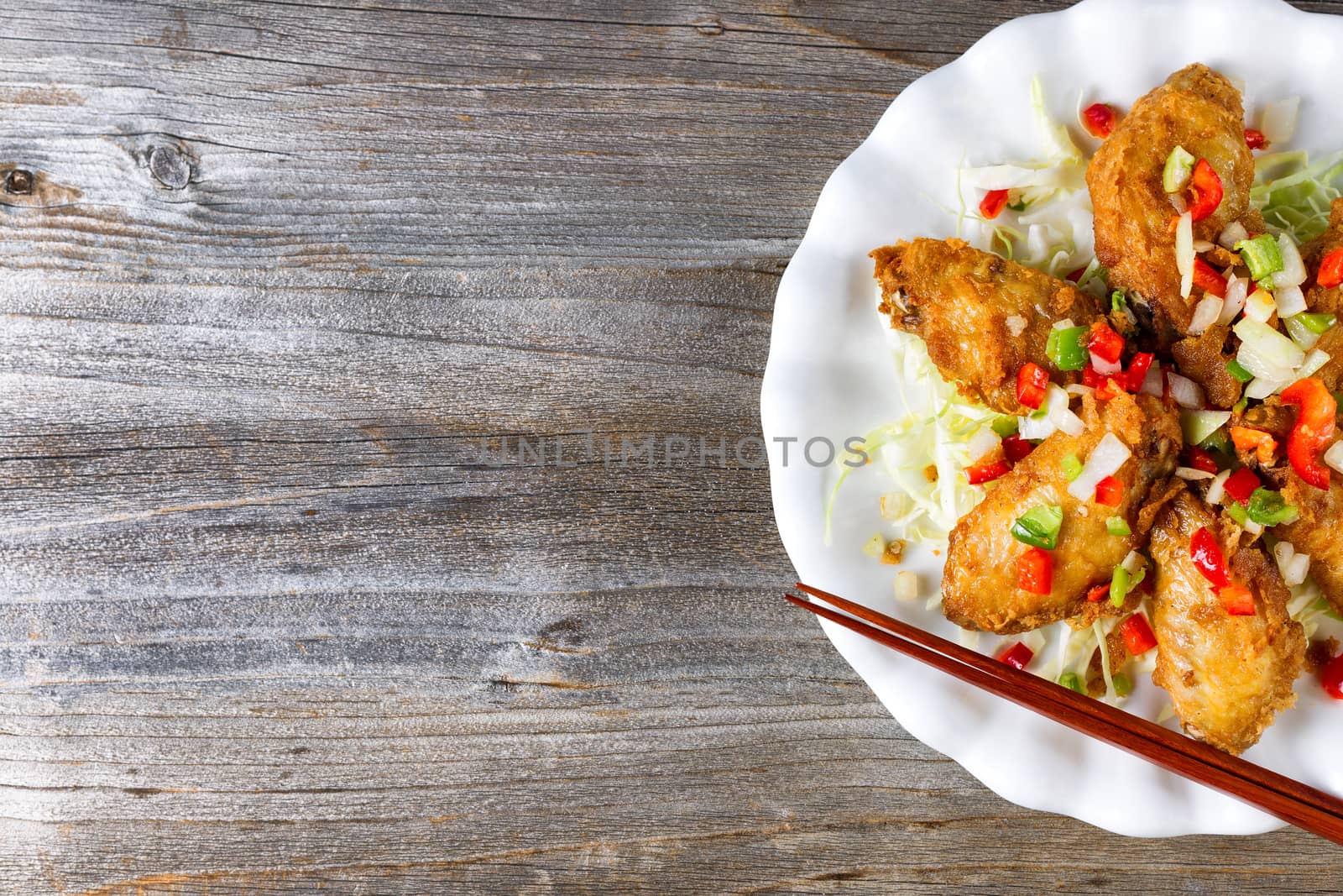 Top view of a partial fried Asian style chicken wings in white plate with garnishes. Rustic wood underneath with copy space on left side.  