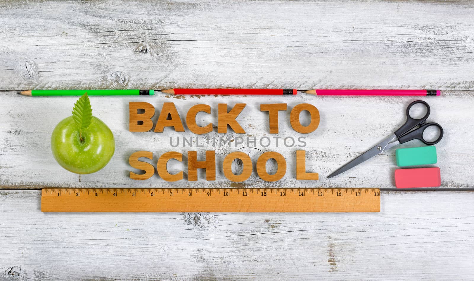 Wooden letters spelling back to school with green apple, pencils, erasers, ruler and scissors on top of rustic white boards.