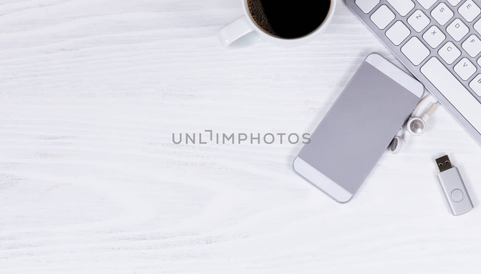 Overhead view of white desktop with partial computer keyboard, cell phone, coffee, thumb drive, and ear phone. 