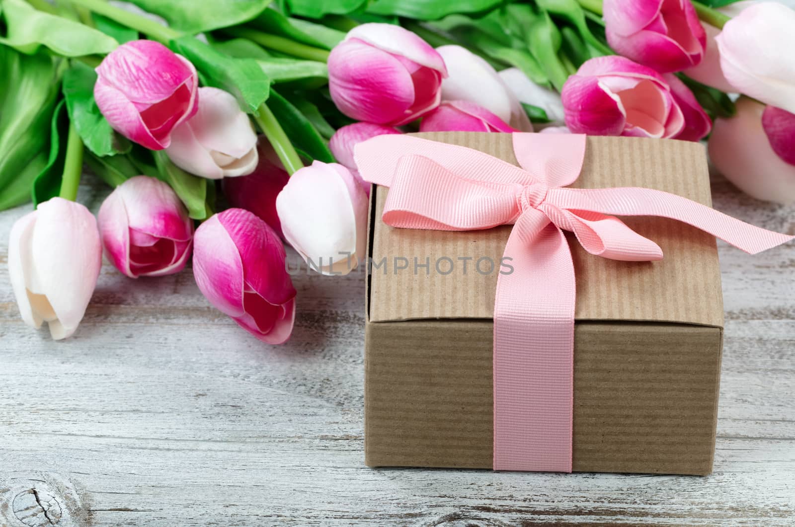 Close up view of a brown gift box with pink tulips in background on white weathered wooden boards 