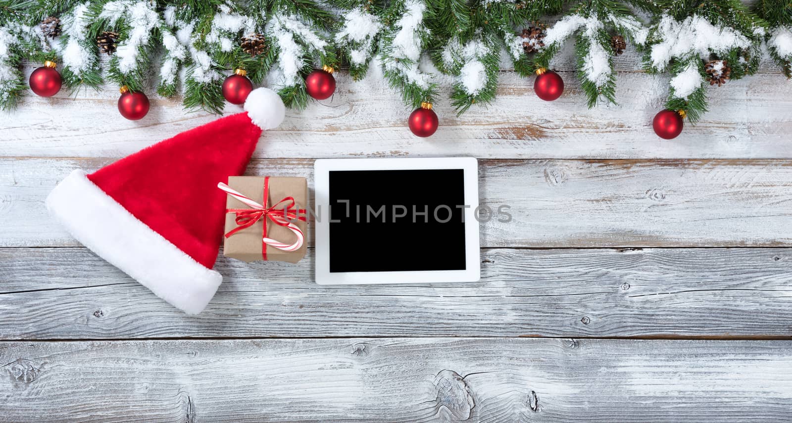 Christmas background with Santa Claus hat, electronic notebook and gift with traditional decorations on rustic white wood. 