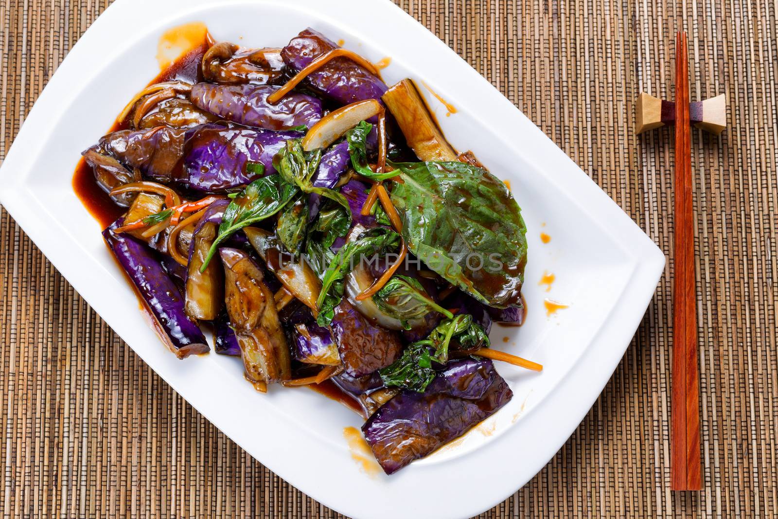 High angled view of cooked eggplant, onion and basil leaves in juicy sauce. Chopsticks in holder with bamboo mat underneath. 