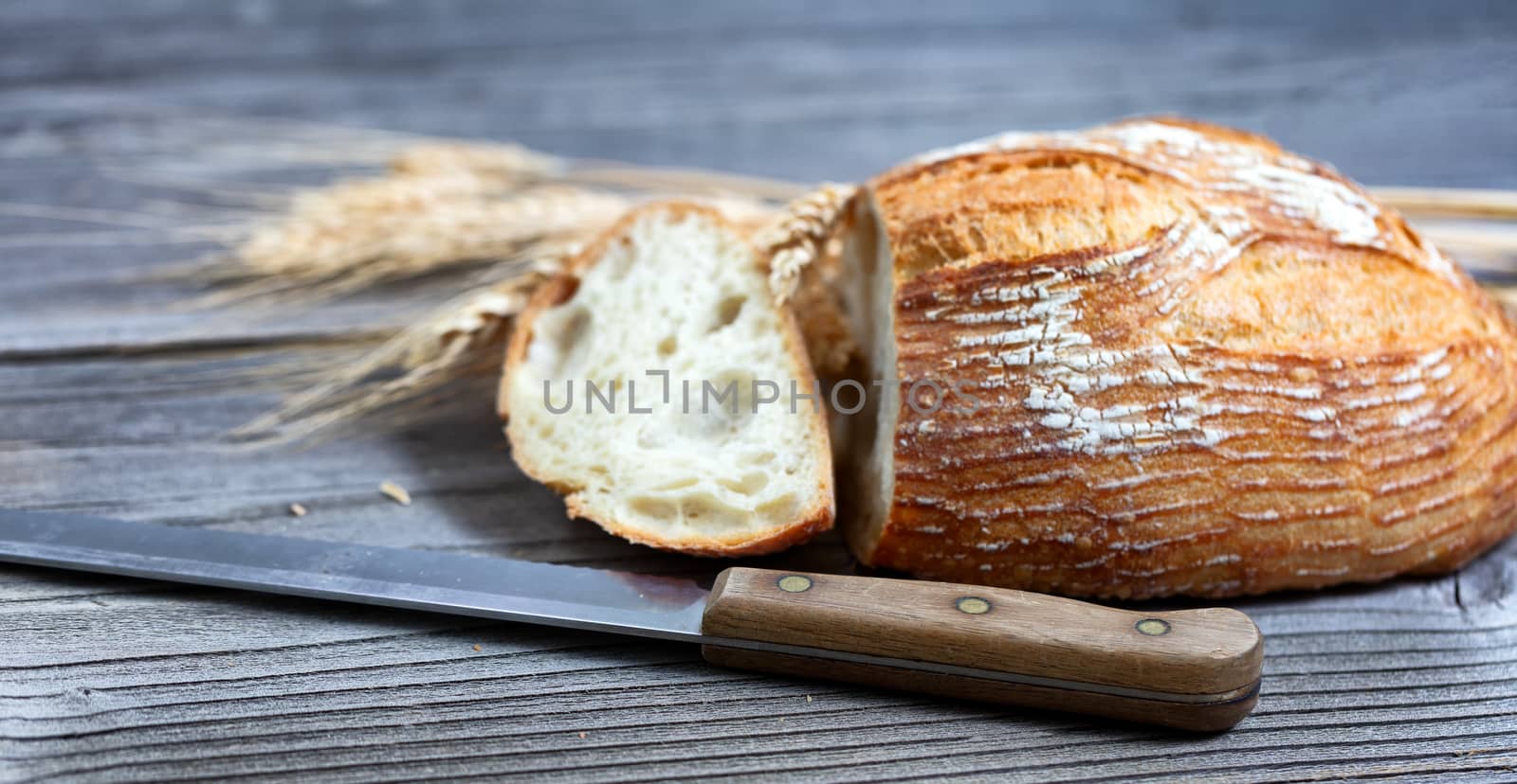 Close up view of a sliced baked whole loaf of bread with cutting knife and wheat stalks on rustic wooden boards