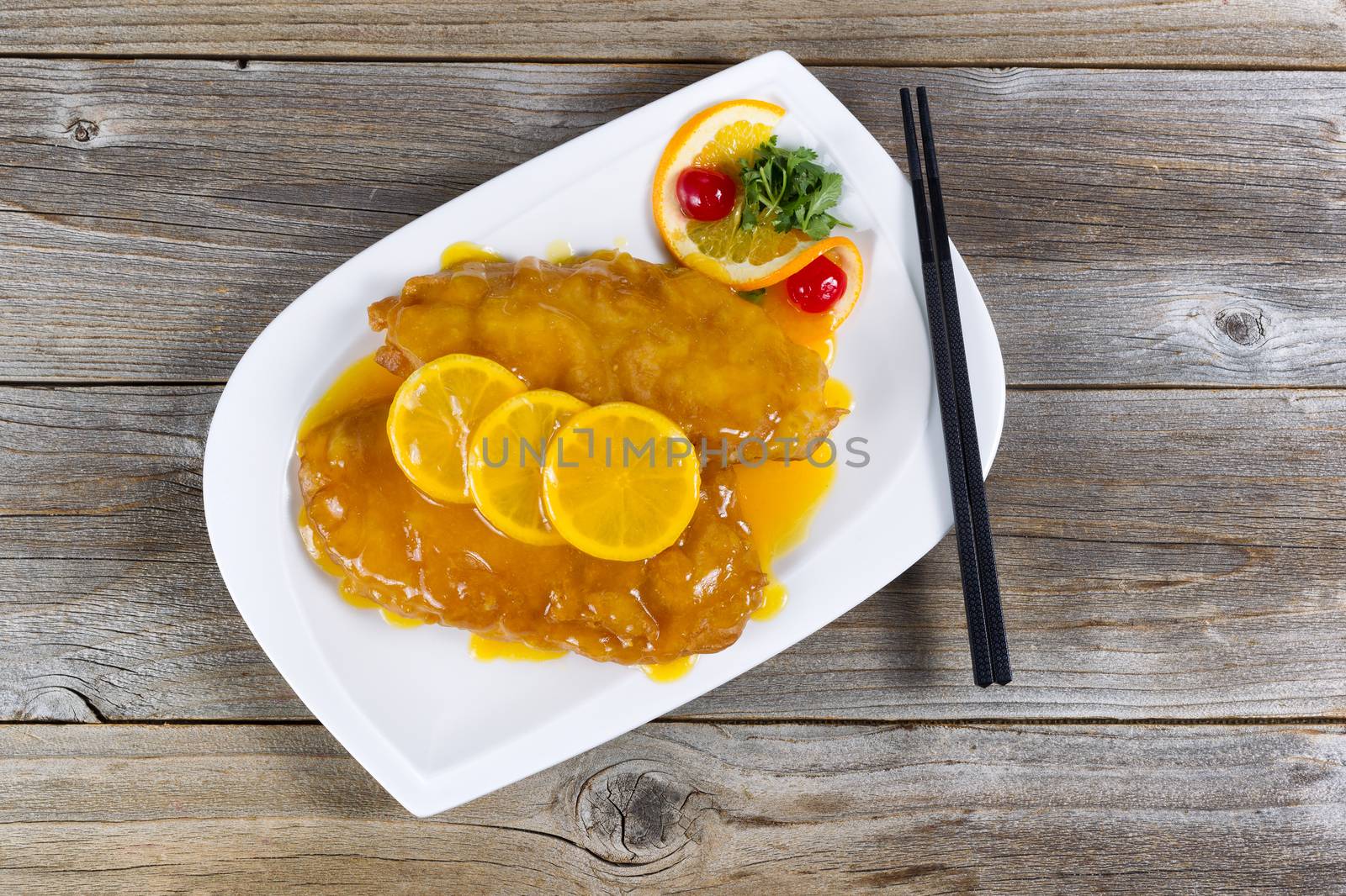 Top view of lemon chicken with tangy sauce and slice lemons on top in white plate with rustic wood underneath.  