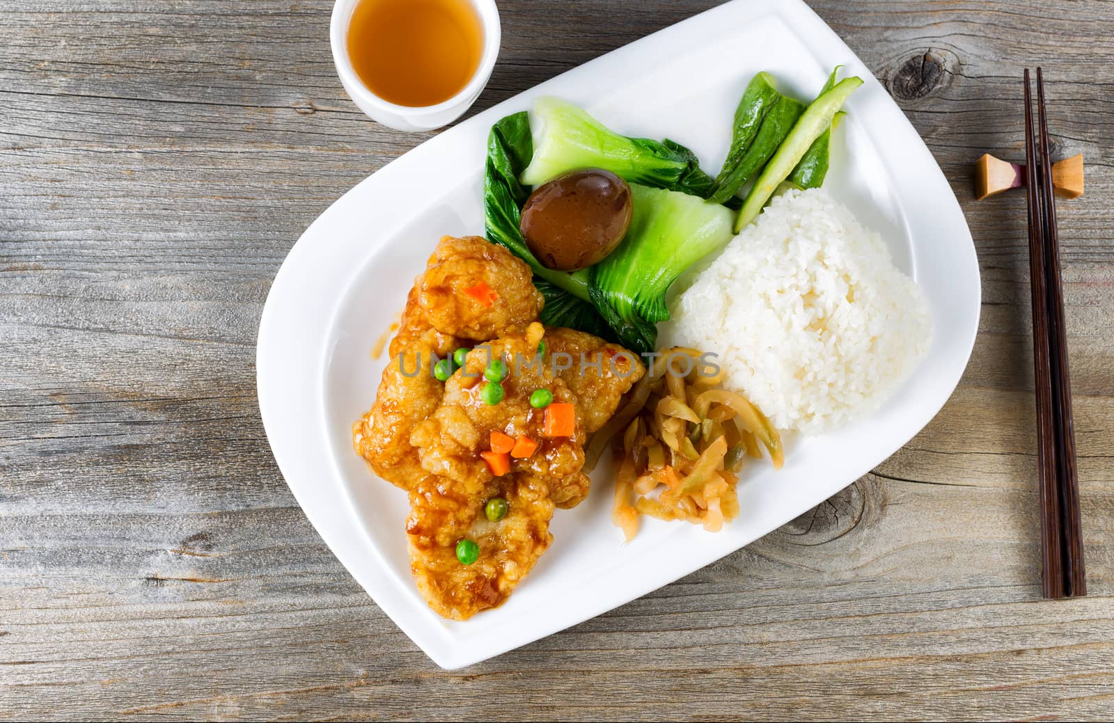 High angled view of lunch consisting of fried bread coated fish, bok choy, rice, egg, and on white plate. Chopsticks and green tea in background. 