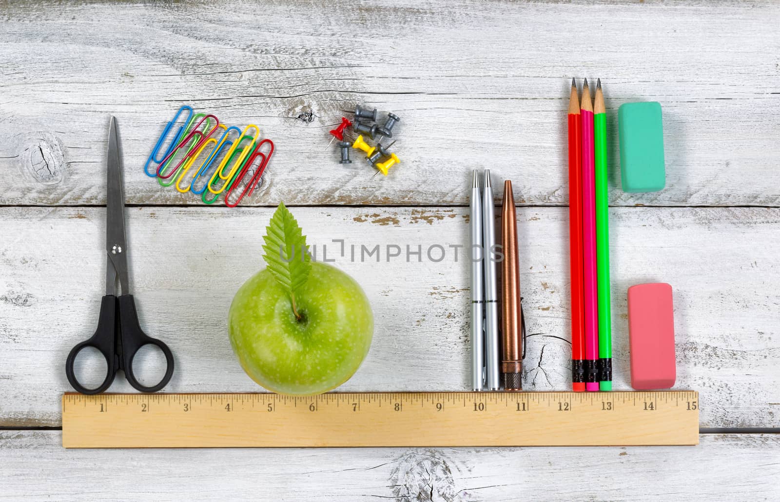 Green apple, pens, pencils, erasers, paper clips, tacks and scissors lined up with ruler on top of white desktop. Education concept. 