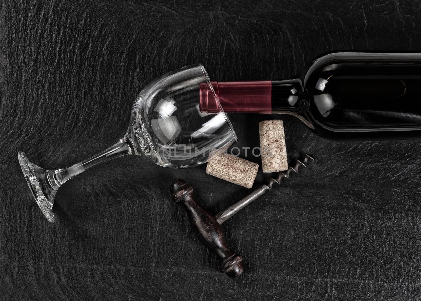 Overhead view of antique wine corkscrew, red wine bottle, glass and used corks on black slate 