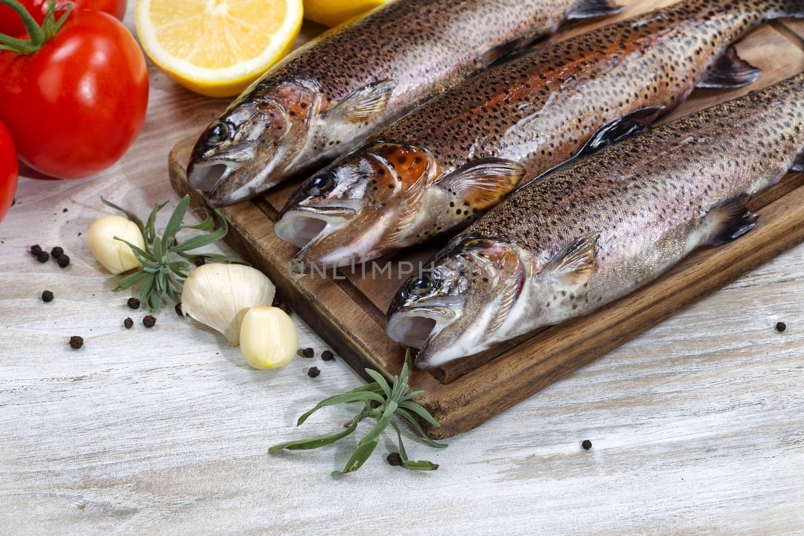 Close up of fresh wild trout being prepared, skin coated with oil, for cooking on server board. Herbs, tomatoes, lemon and spices on white aged wooden boards. 