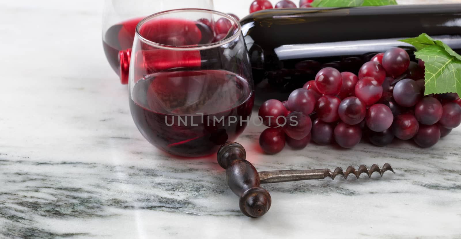 Close up view of a corkscrew with red wine bottle, filled glasses and grapes on natural marble stone setting in background