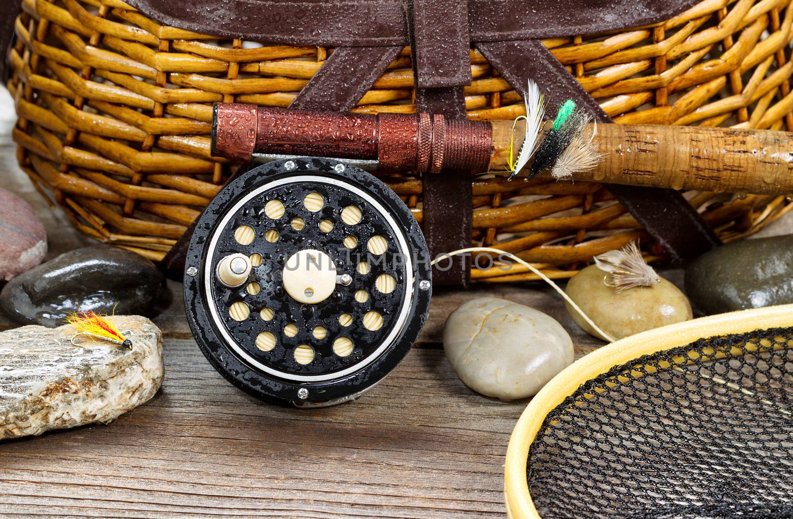 Close up of a wet antique fly fishing reel, rod, landing net, artificial flies and rocks in front of creel with rustic wood underneath. Layout in horizontal format.