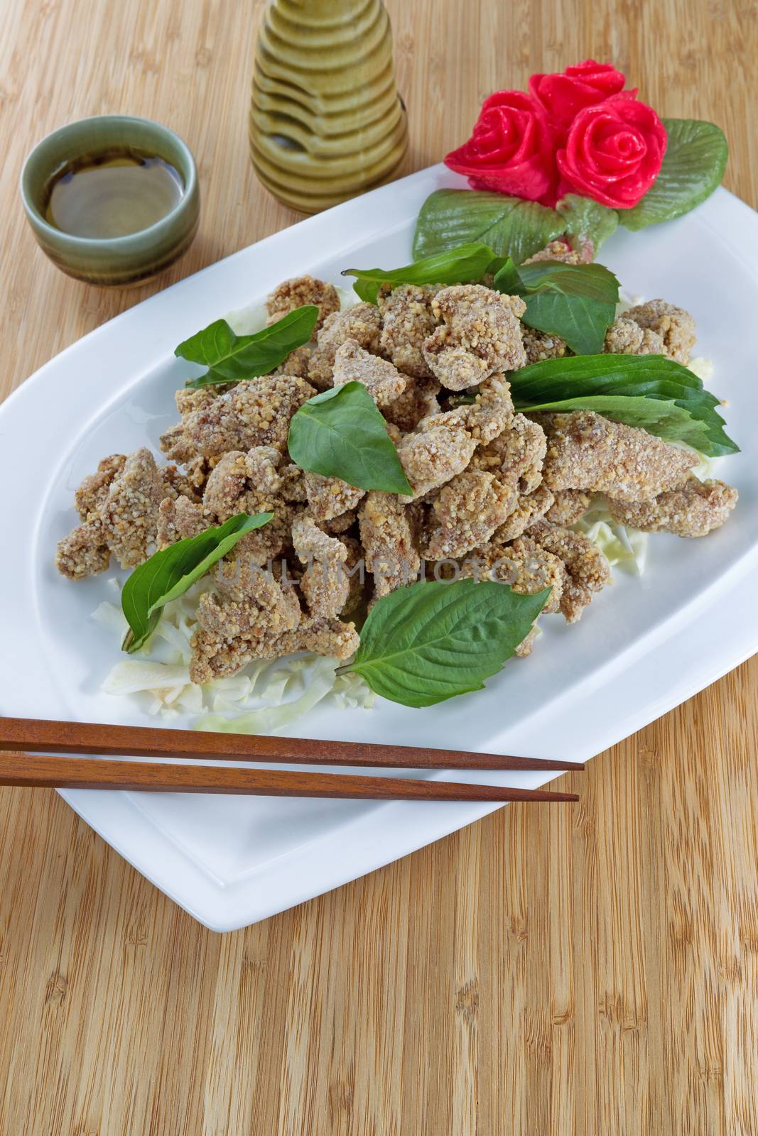 Vertical image of Chinese sliced fried cooked chicken in white dish with cup and ceramic drink pitcher in background on natural bamboo background 