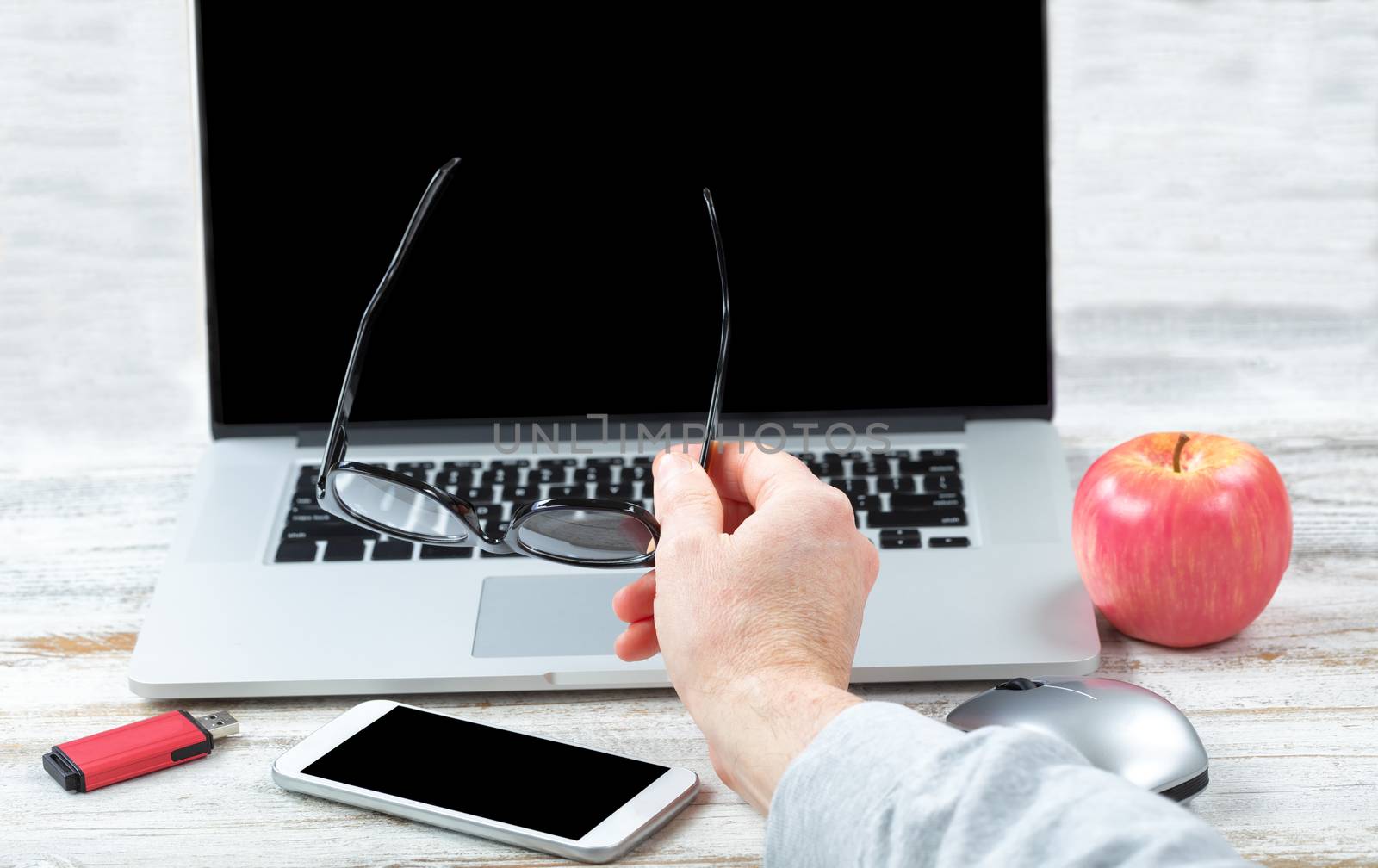 Close up of male hand holding reading glasses with workstation technology in background for business or education use 