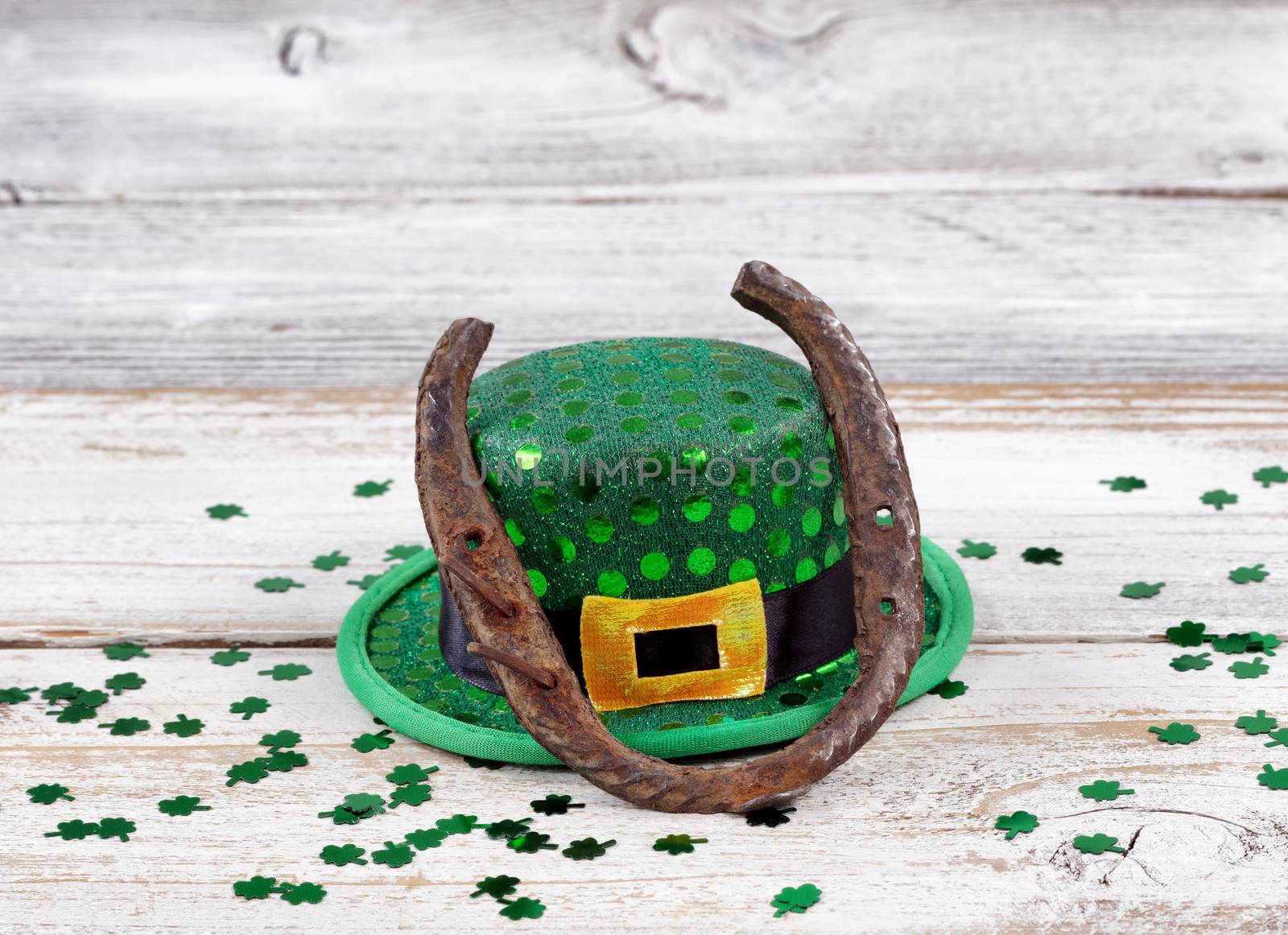 Close up view of a rusty horseshoe resting on hat for St Patrick day with shiny clovers on weathered white wooden boards 