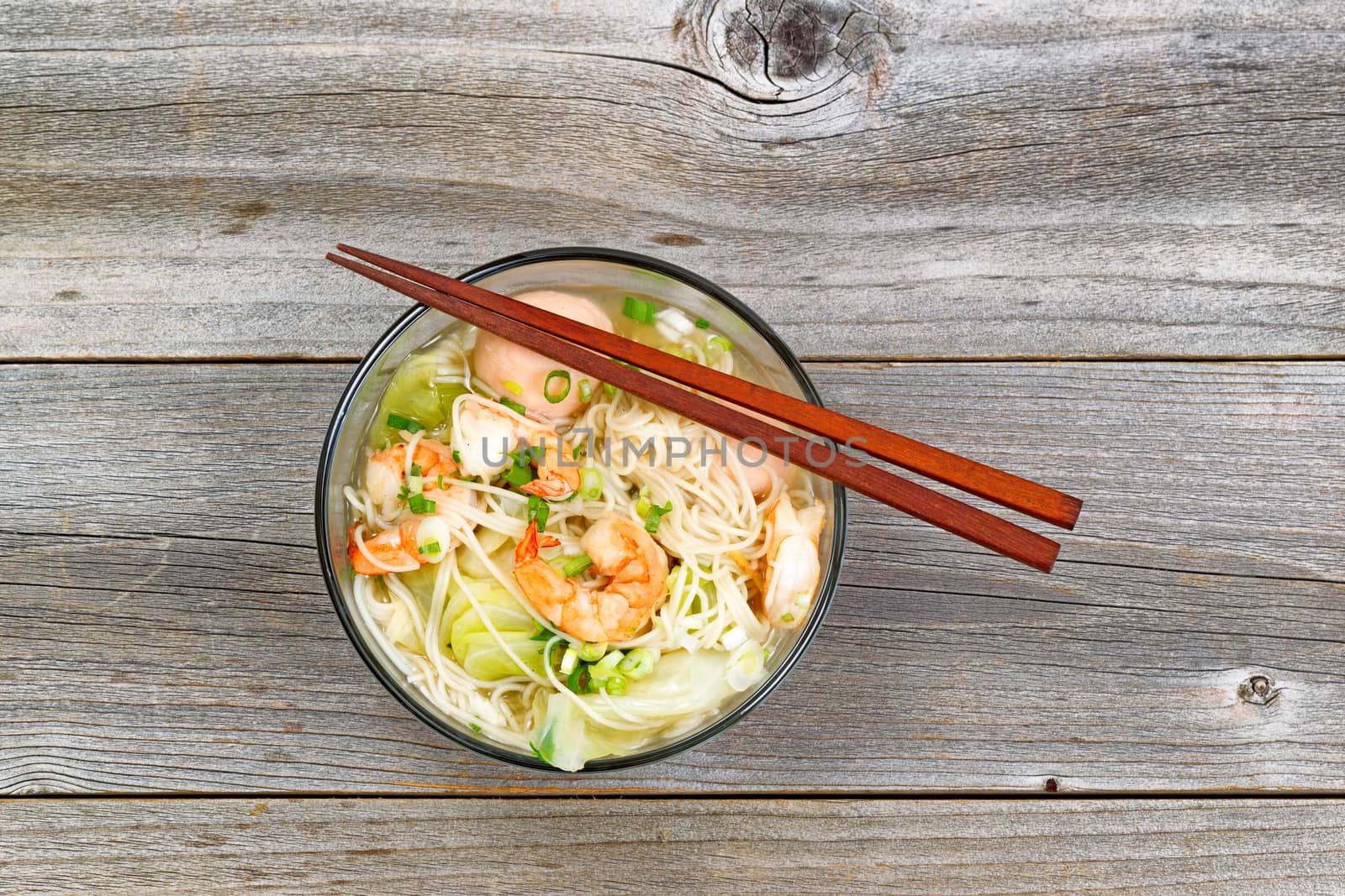 Top view image of Chinese noodle soup in clear glass bowl on top of rustic wood. Layout in horizontal format.