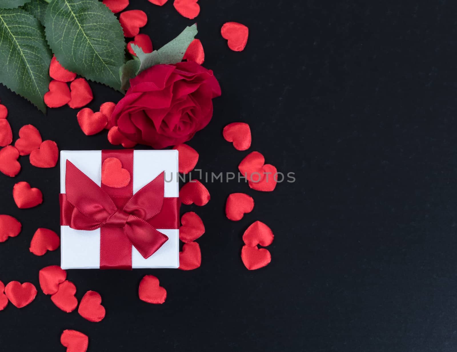 Overhead view of a gift box, heart shapes and a single red rose on dark stone background