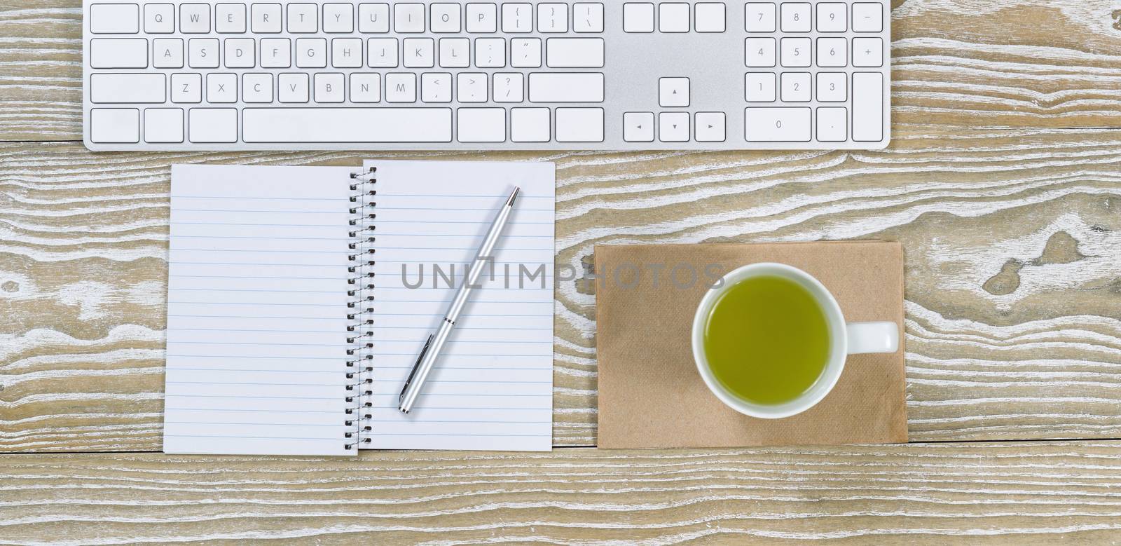 Top view shot of an old white desktop with keyboard, green tea in cup, notepad and pen in horizontal format.