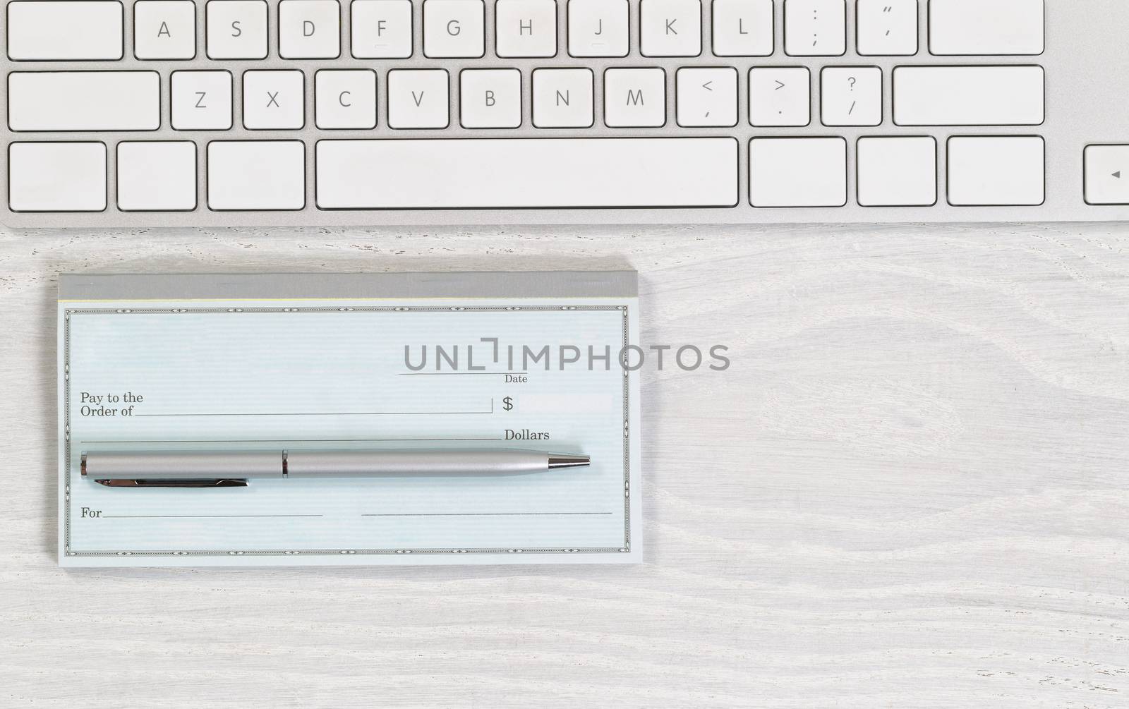 Image of partial keyboard with blank checkbook and silver pen on white desktop. Layout in horizontal format.