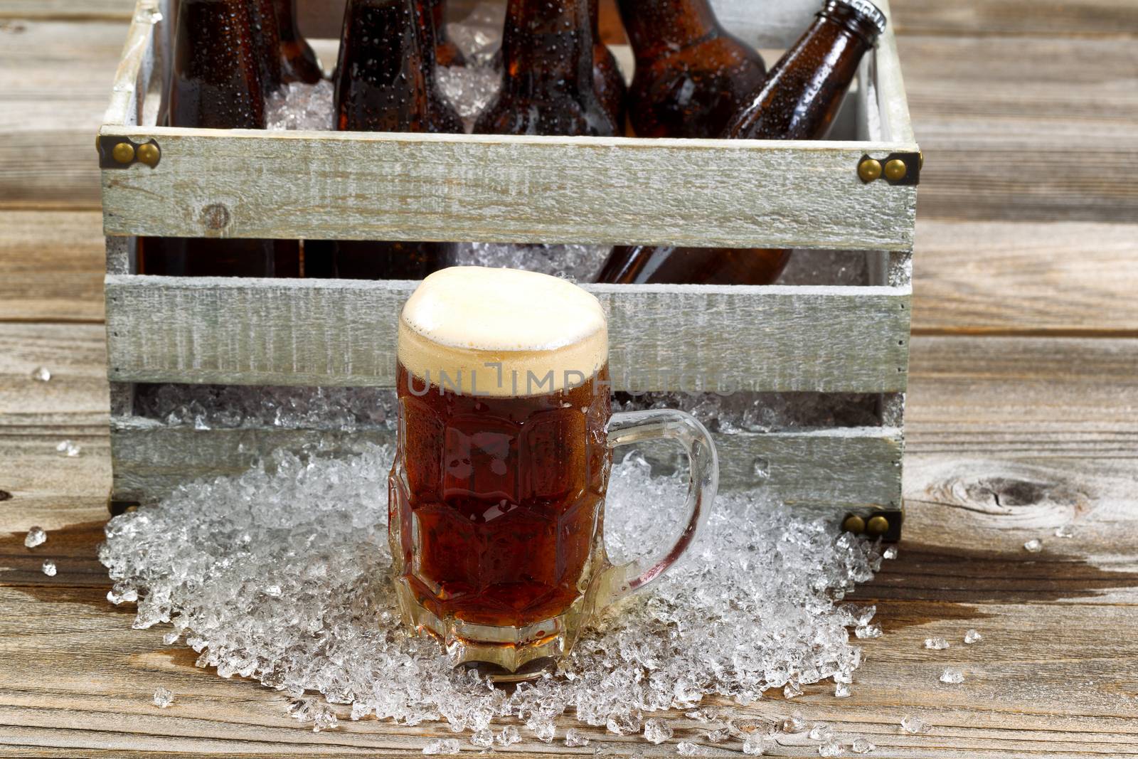 Frosty glass mug of dark beer, focus on front, with vintage crate filled with bottled beer and crushed ice on rustic wooden boards. 