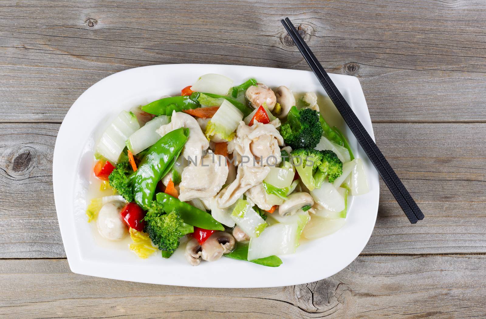 Top view of stir fried white chicken pieces with broccoli, snow peas, peppers and mushroom in white plate on rustic wood setting. 