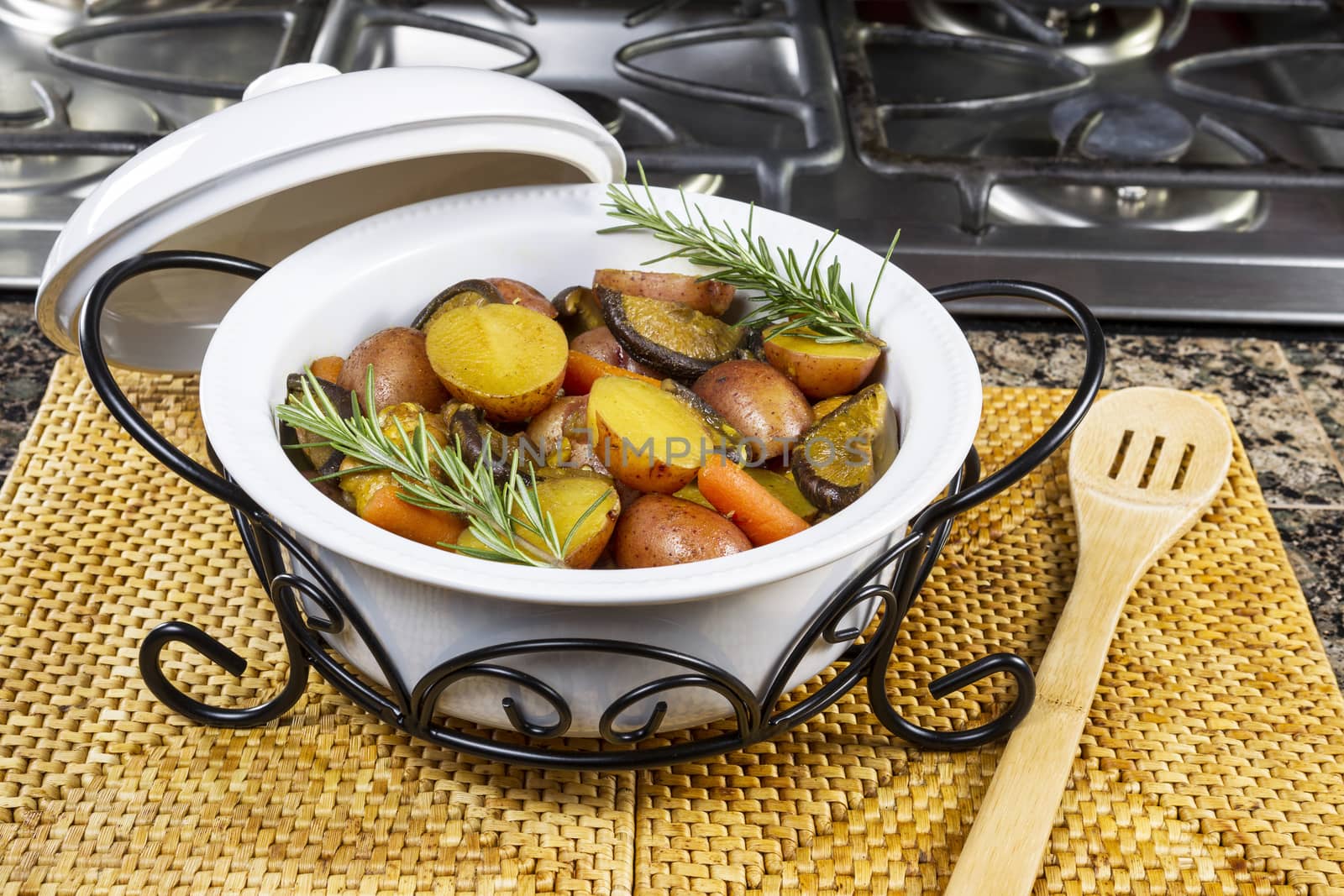 Fresh pot of curry chicken, potatoes, mushroom and carrots on top of place mat with wooden spoon next to stove top range in kitchen  