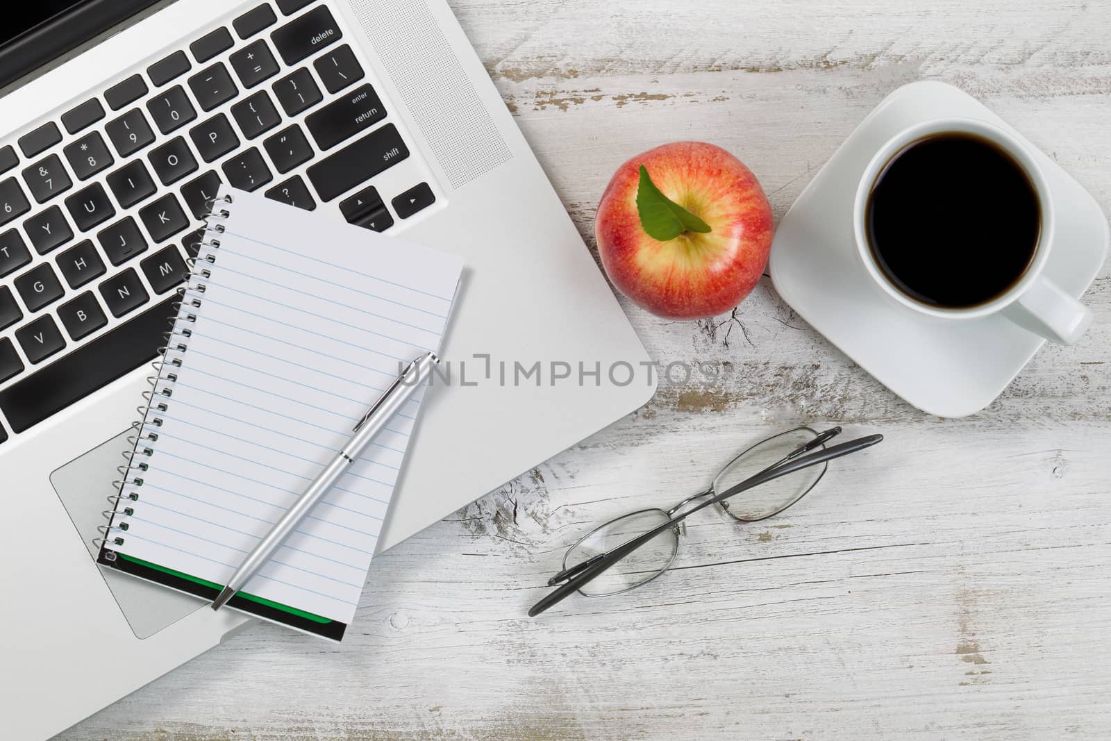 Top view of desktop with partial laptop, apple, coffee, reading glasses, and pen. 