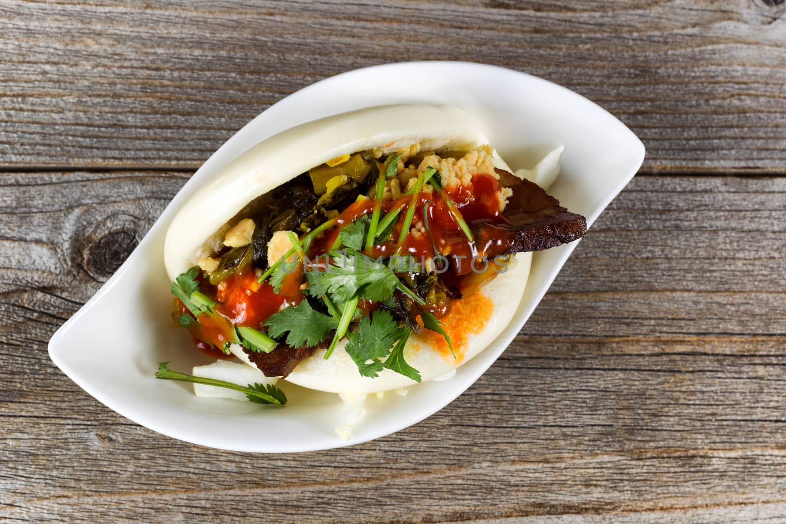 Top view of a slice of beef meat in bun with sauce and herbs inside small white bowl on rustic wood. 