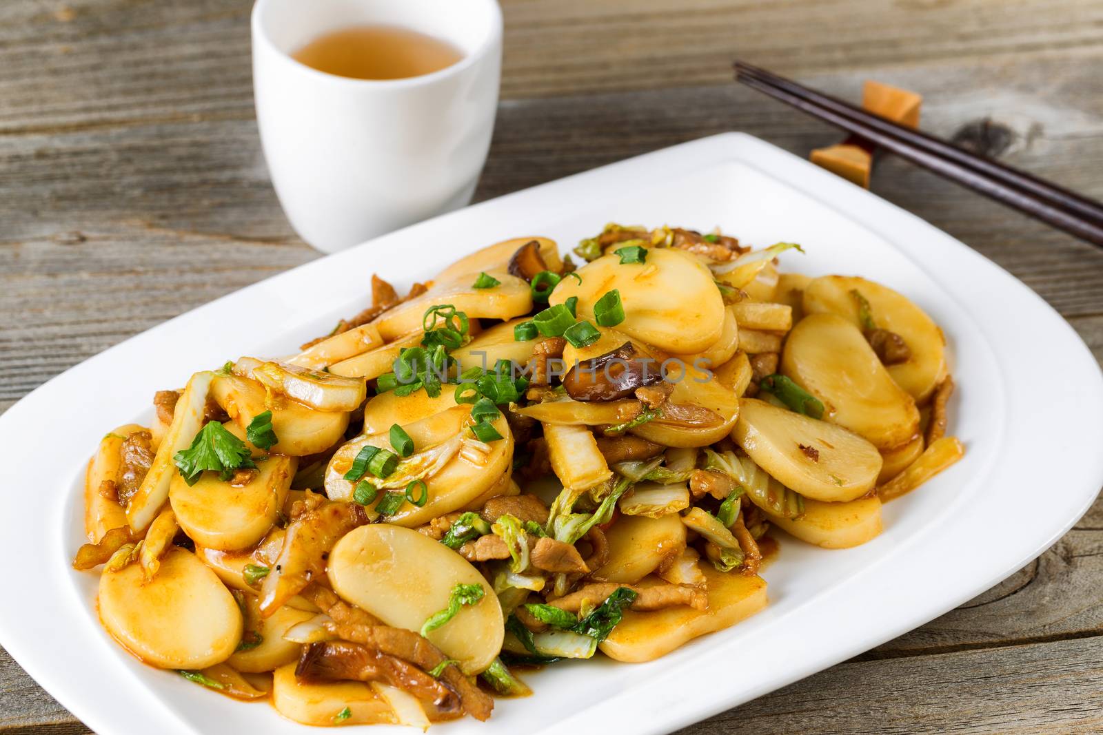 Close up front view of stir fry consisting of sliced sticky rice, onion, mushroom, and chicken. Chopsticks and tea in background on rustic wood. Selective focus on front of dish. 
