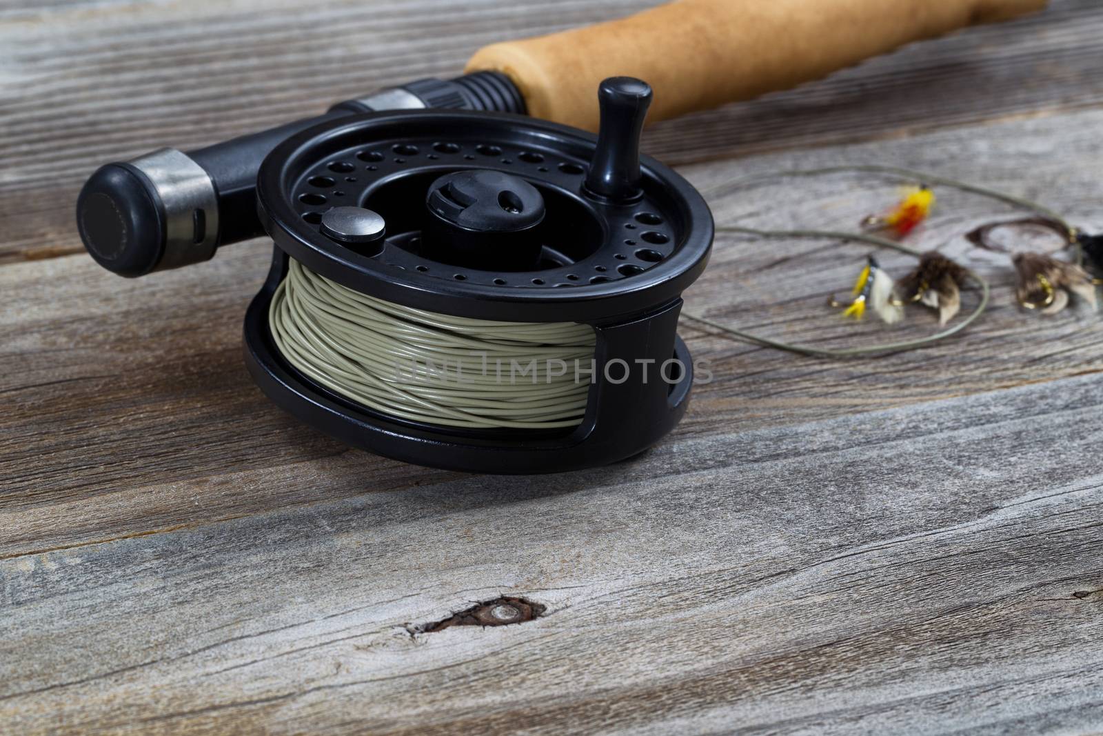 Close up of fly reel, focus on front bottom of reel, with partial cork handled pole and flies blurred out on rustic wooden boards 

