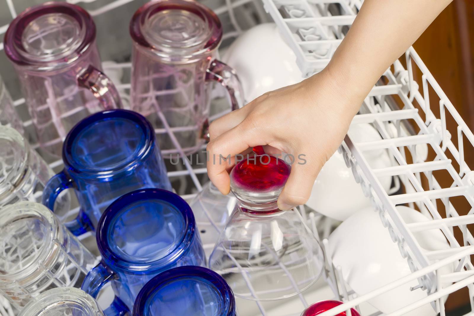 Horizontal photo of female hand putting in glassware to be washed by dishwasher with blue, red, clear, pink glasses and white bowls in background