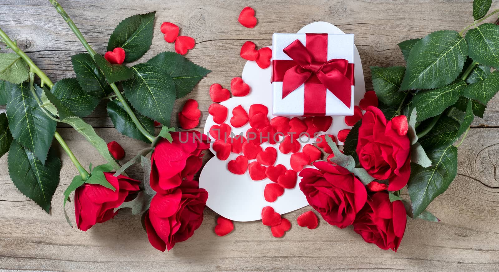 red roses surrounding white gift box and many hearts on weathered wooden background in flat lay view 