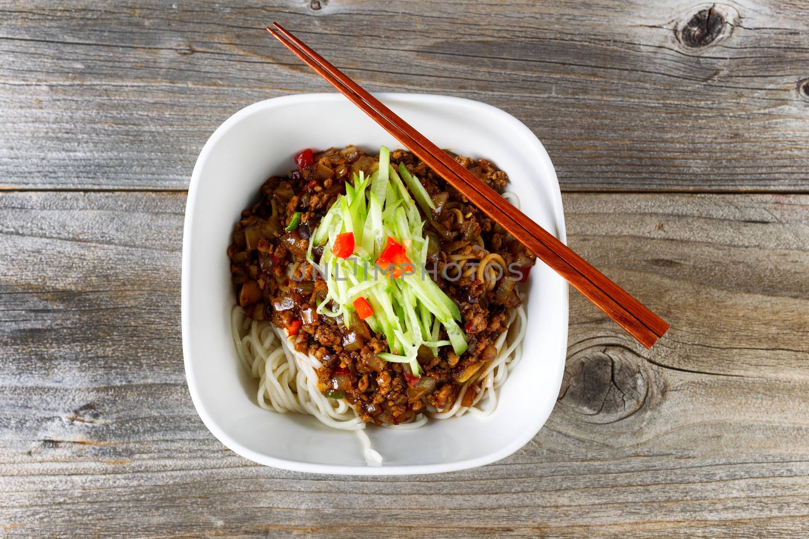 High angled view of noodles with ground beef and cucumbers. Chopsticks on top of bowl with rustic wood underneath. 