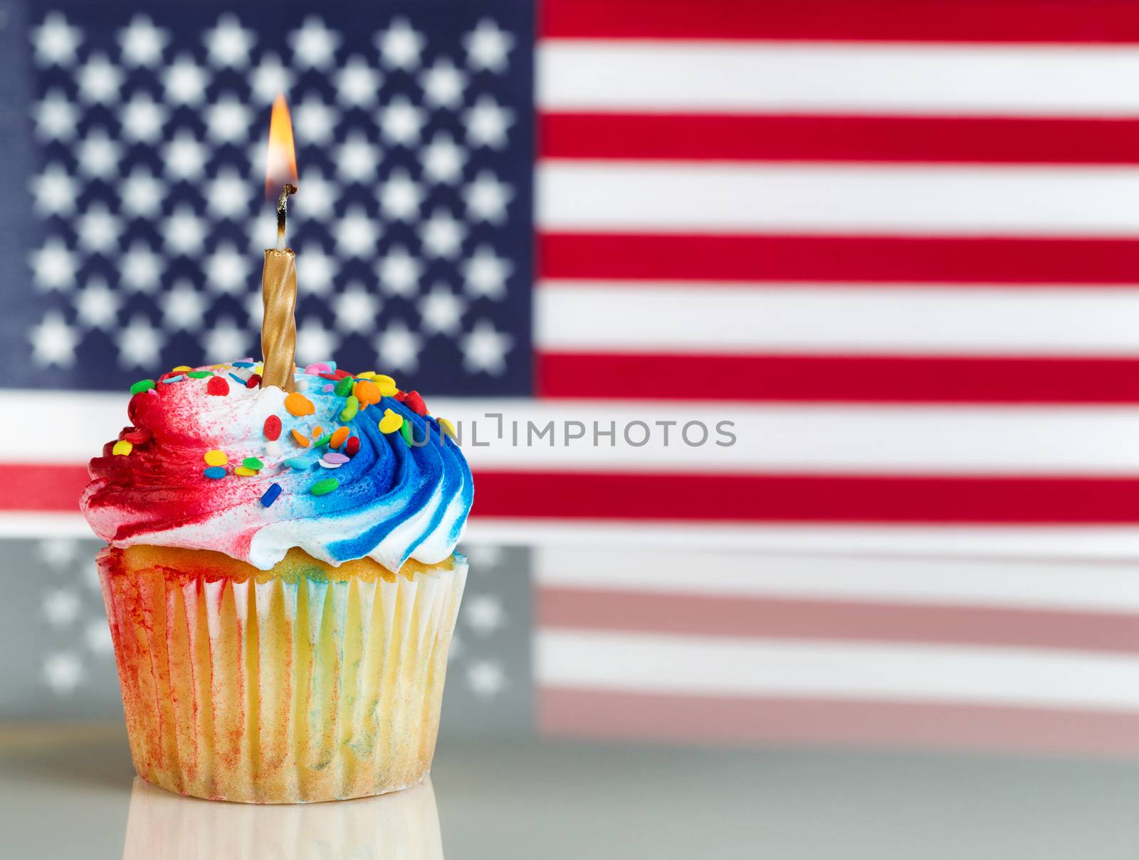 Close up of a red, white, and blue frosted cupcake with burning candle. Fourth of July concept with United States of American flag in background.
