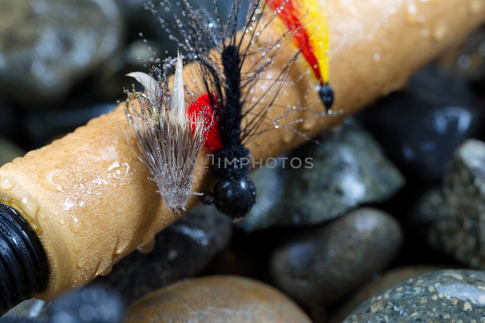 Close up of  trout flies, focus on front one with shallow depth of field, on wet cork handle of fishing rod with blurred out river rocks in background