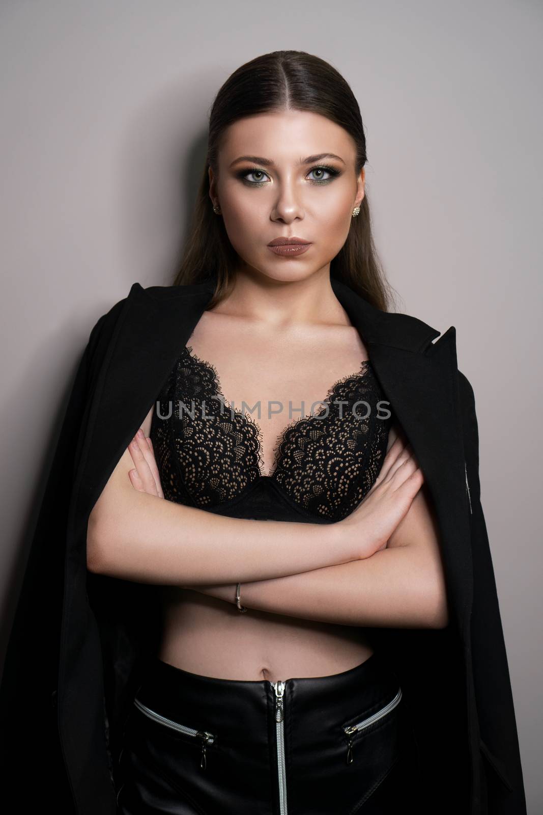 Beautiful young white girl close-up in a black jacket on a gray background. Make-up artist, beauty salon, magazine. Arms crossed. Vertical portrait.
