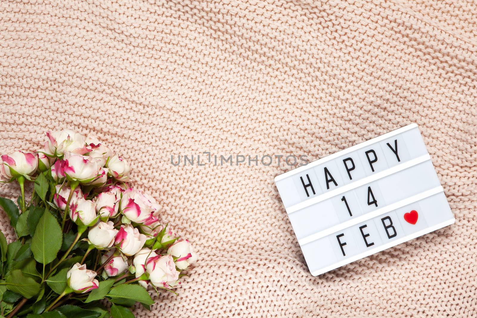 Bouquet of small roses on dusty pink blanket, lightbox with inscription Happy 14 FEB that means Valentine's Day. Flat lay. Top view. February 14th celebration concept. Horizontal, copy space.