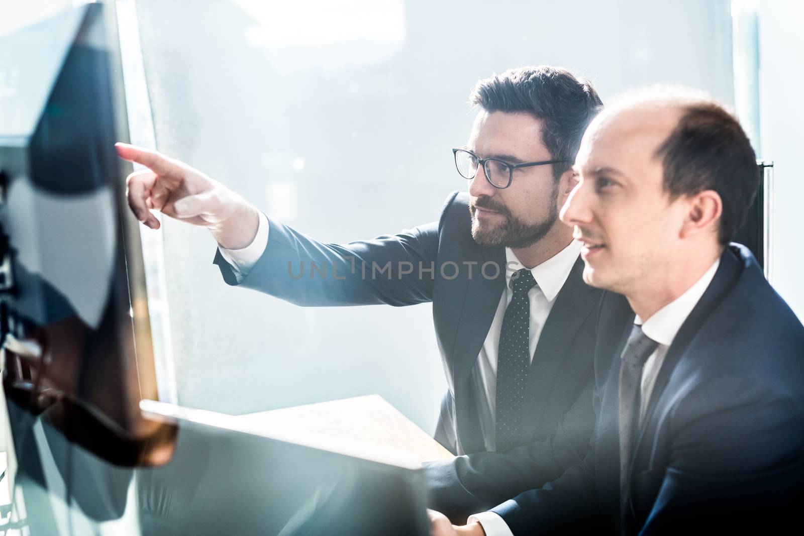 Image of two thoughtful businessmen looking at data on multiple computer screens, solving business issue at business meeting in modern corporate office. Business success concept.